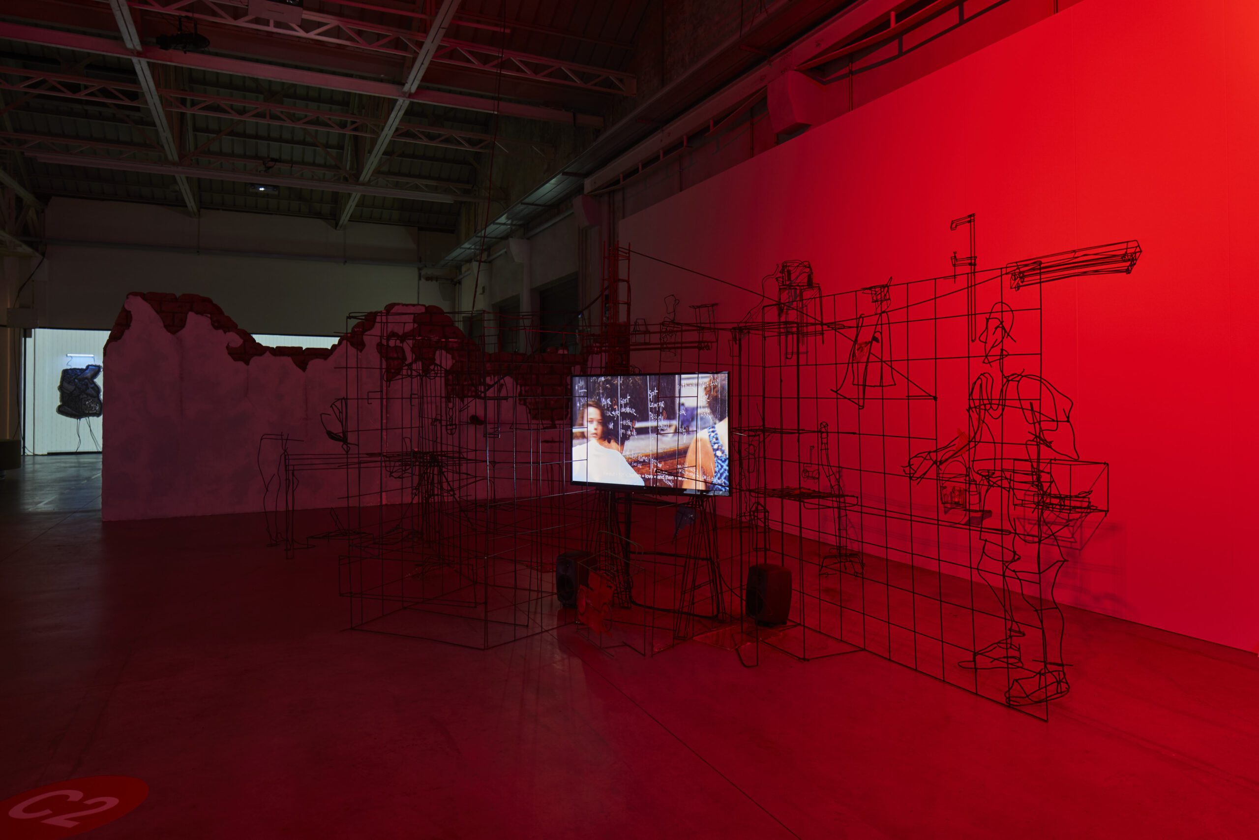 Un'installazione artistica con una luce rossa fioca, figure in filo di metallo e uno schermo video su scena urbana—un'arte per impresa in uno spazio industriale.
