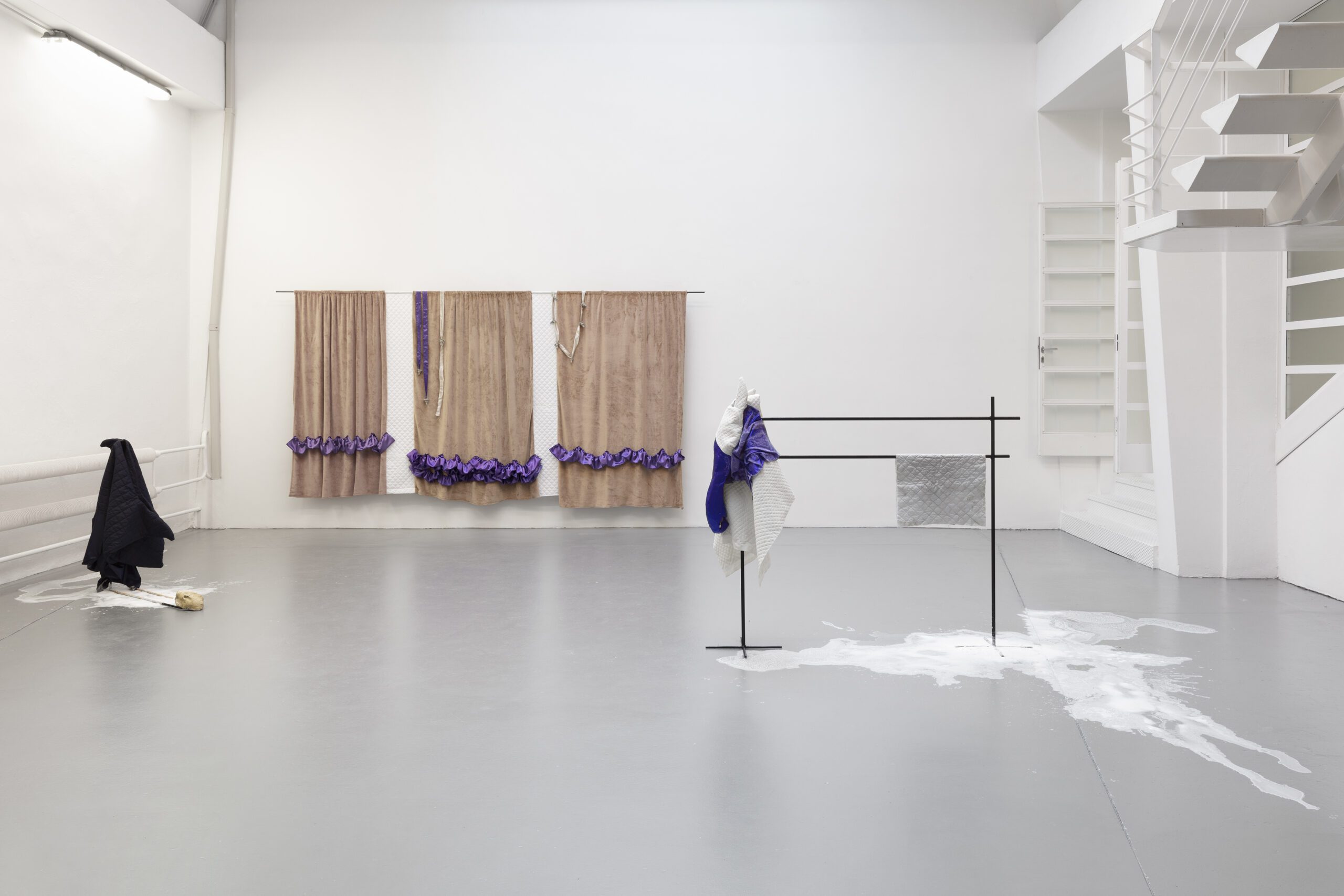 Galleria minimalista: tessuti sospesi, una struttura nera, liquido bianco sul pavimento e una scala che ispira eventi ed esperienze.